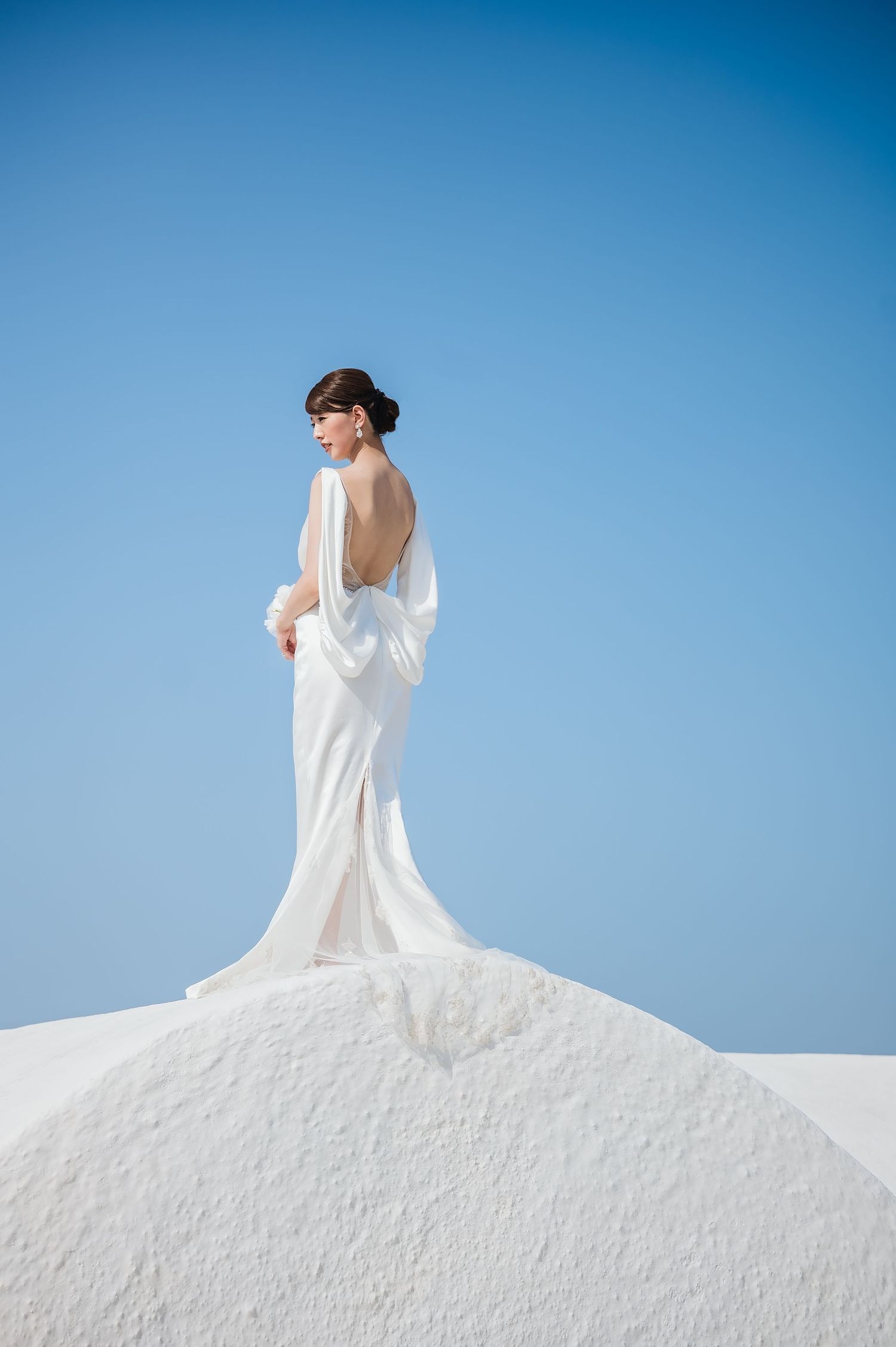 Bride in elegant backless white gown with flowing sleeves stands atop white architecture against clear blue sky.