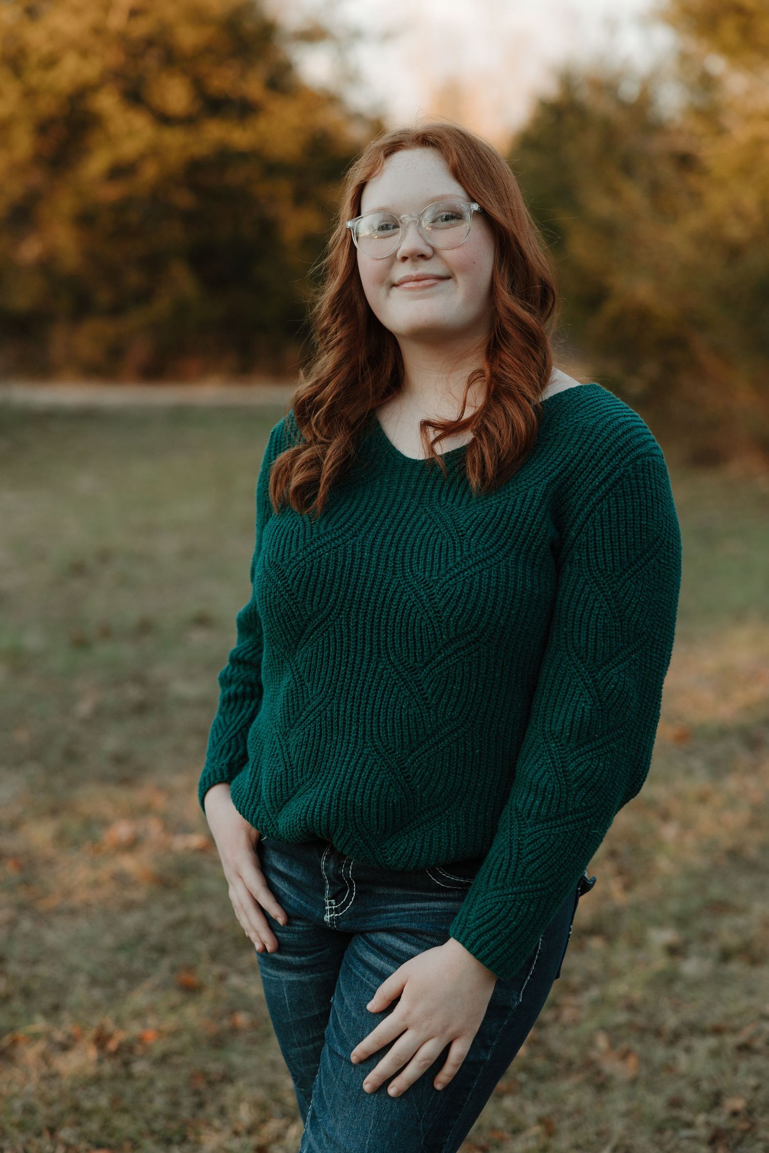 Young woman with red hair and glasses wearing a green knit sweater and jeans, smiling outdoors in autumn setting.