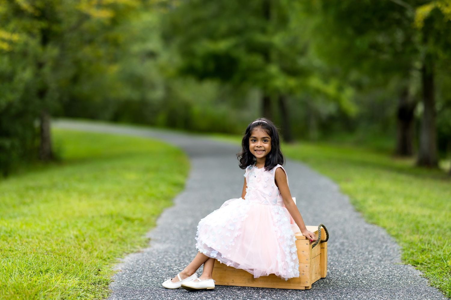 Young girl in pink dress sitting on vintage suitcase on tree-lined path in lush green park setting.