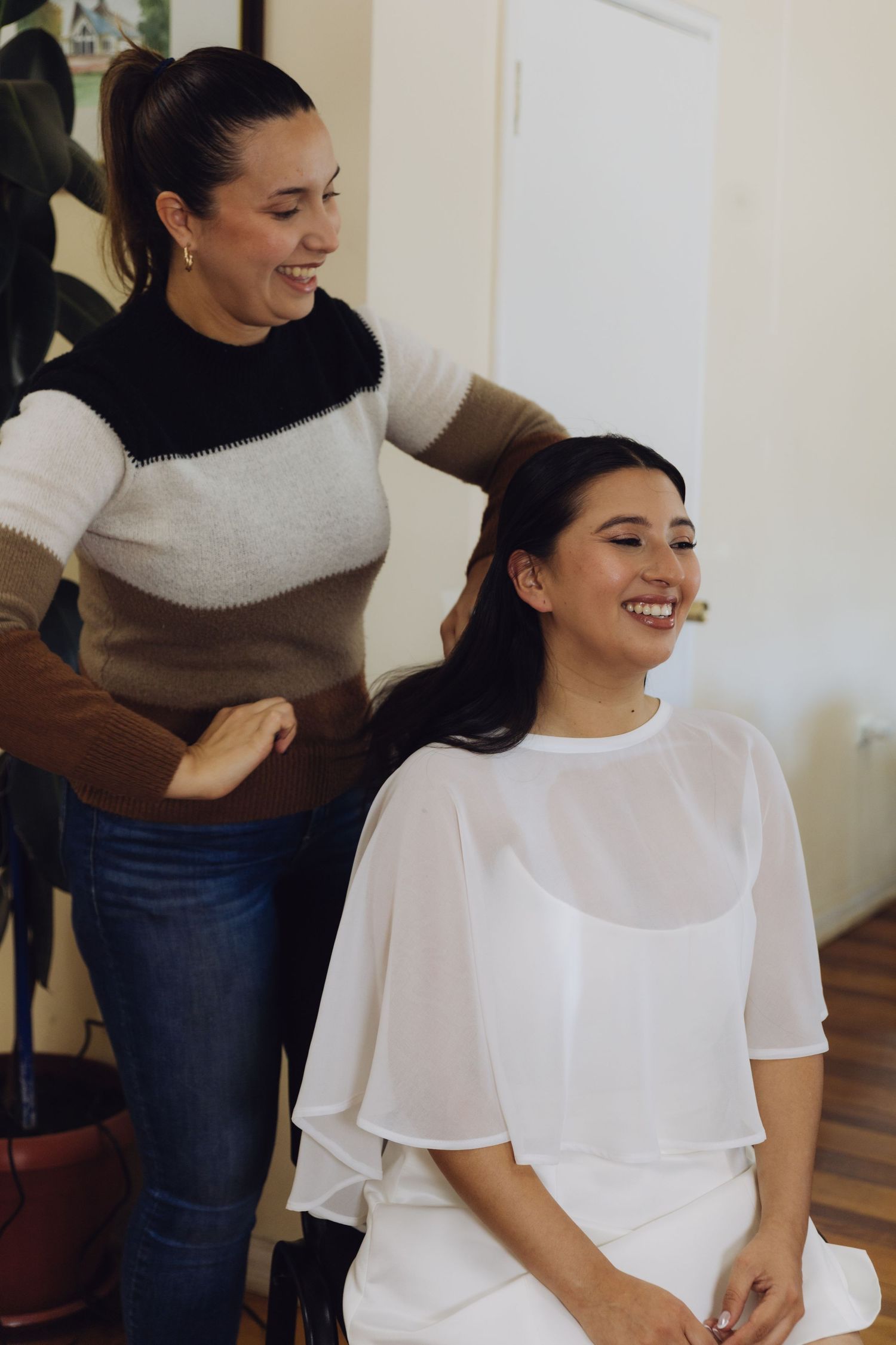 Smiling bride in a white dress laughing as her hairstylist works on her hair before the wedding.