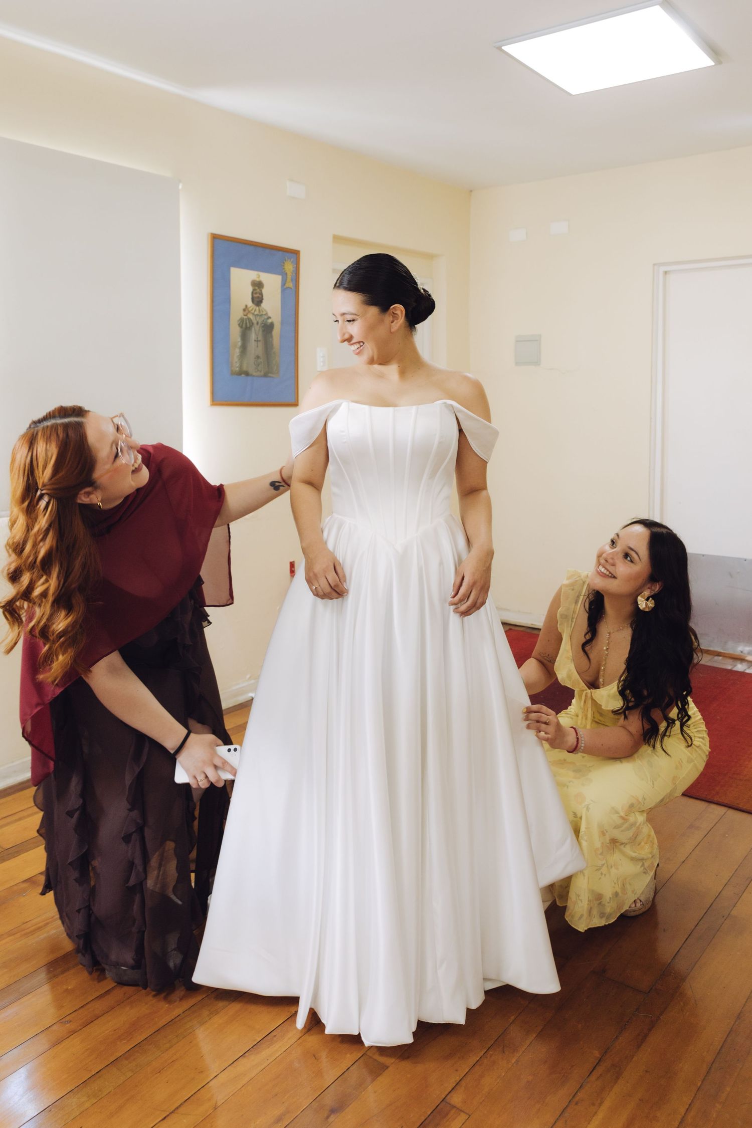 Bride in white off-shoulder gown assisted by two women adjusting her dress indoors.