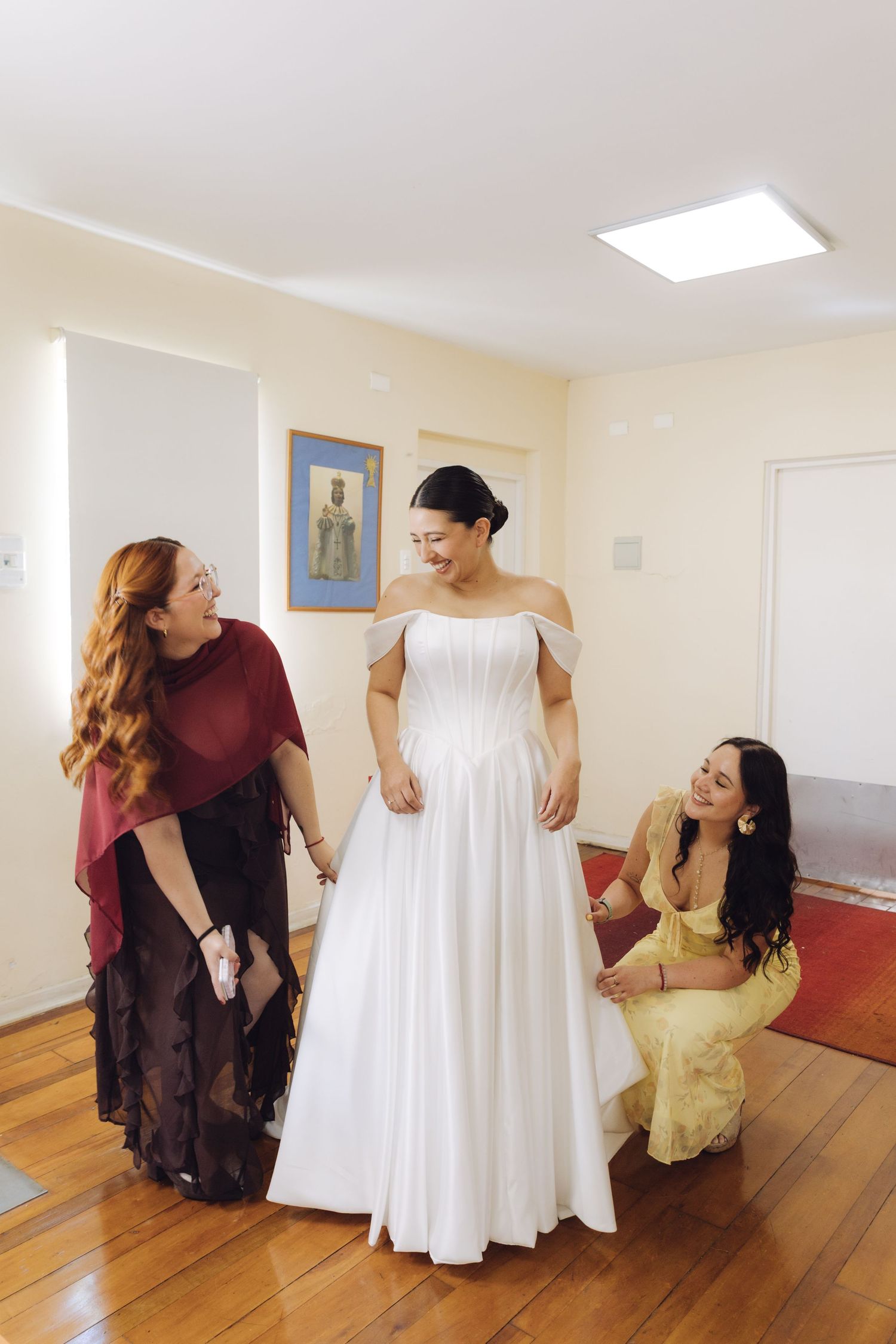 Bride in elegant white wedding gown laughing with two bridesmaids in a warmly lit room.