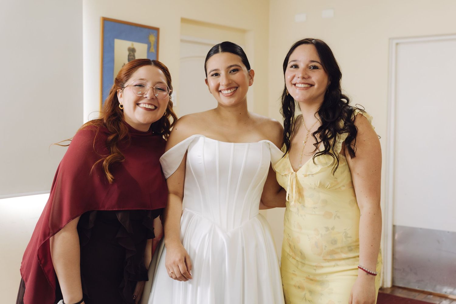 Bride in white strapless gown posing with two smiling women in burgundy and gold dresses.