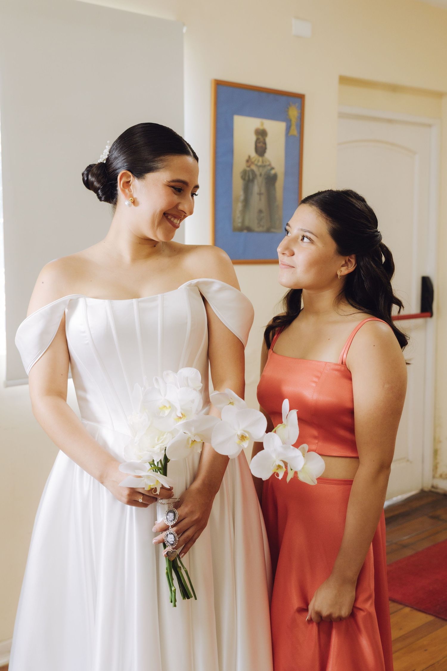 Bride in off-shoulder white gown holding white orchids smiling with bridesmaid in coral dress.