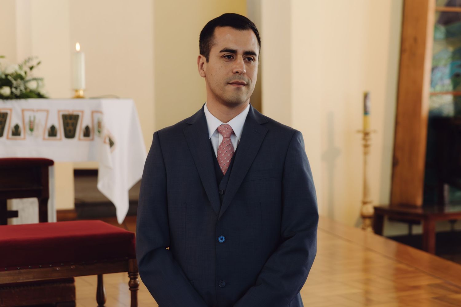 Groom in navy suit with pink tie standing solemnly inside a church before the ceremony.