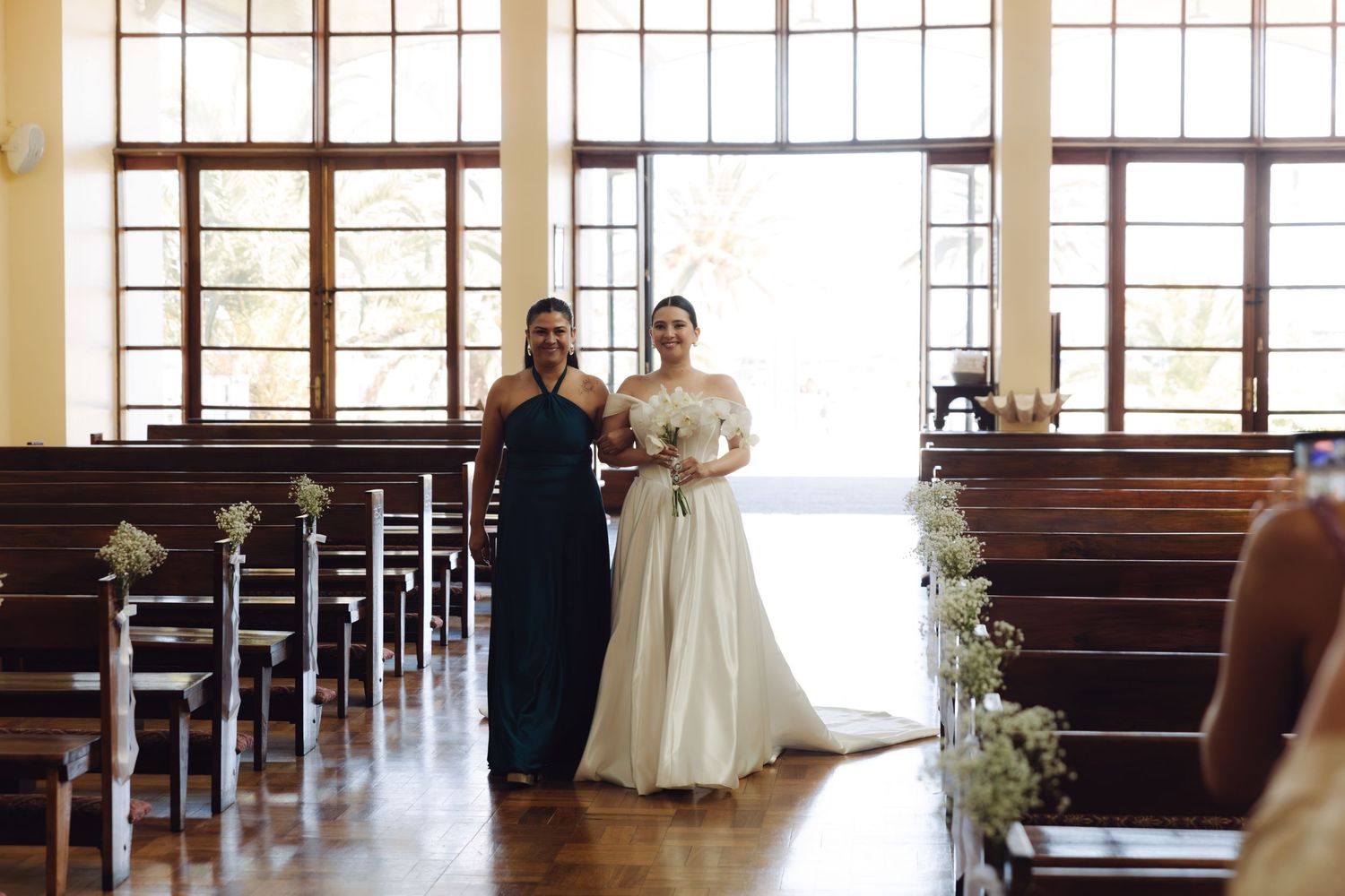 Bride in white off-shoulder gown walking down church aisle with bridesmaid in navy dress.