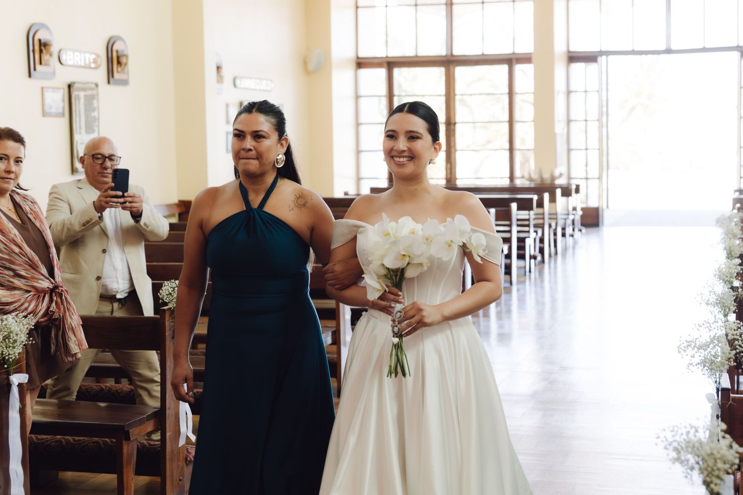 Smiling bride carrying white bouquet walking down church aisle escorted by woman in teal dress.
