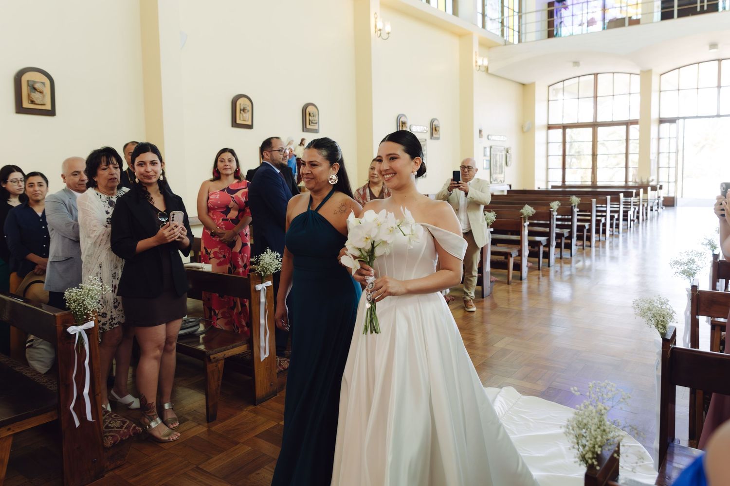 Bride in flowing white gown walking down decorated church aisle surrounded by wedding guests.