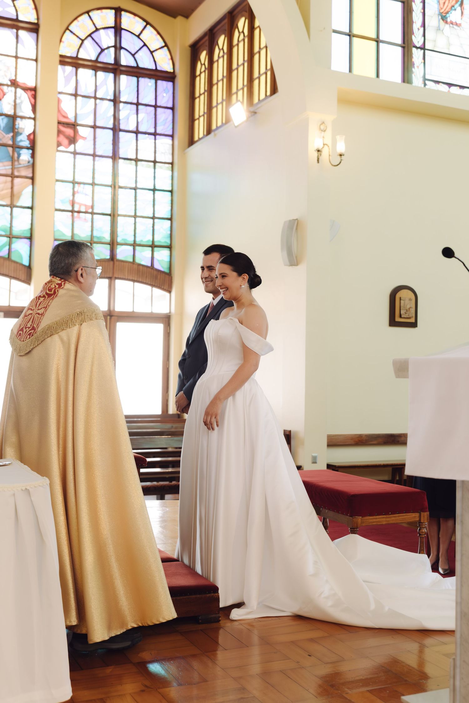 Bride and groom standing before a priest during their wedding ceremony in a sunlit church.