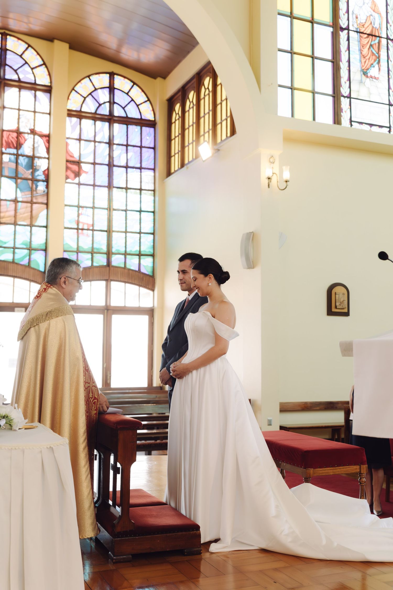 Bride and groom stand before a priest during a church wedding ceremony with colorful stained glass windows.