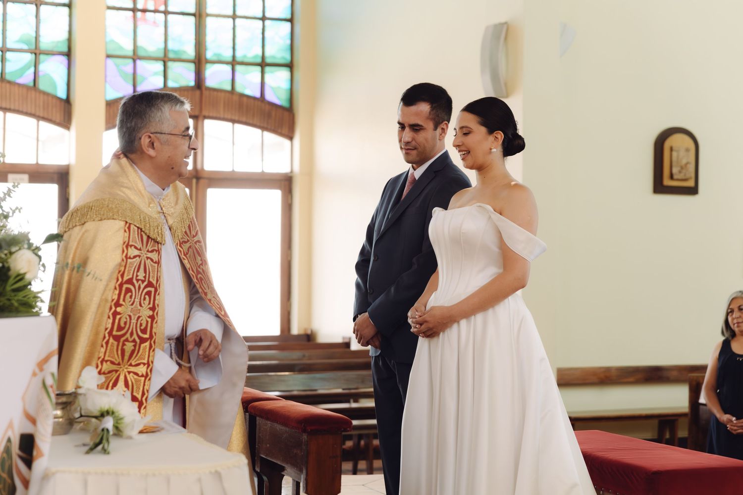 Couple exchanges vows with a priest in golden vestments during an intimate church wedding ceremony.
