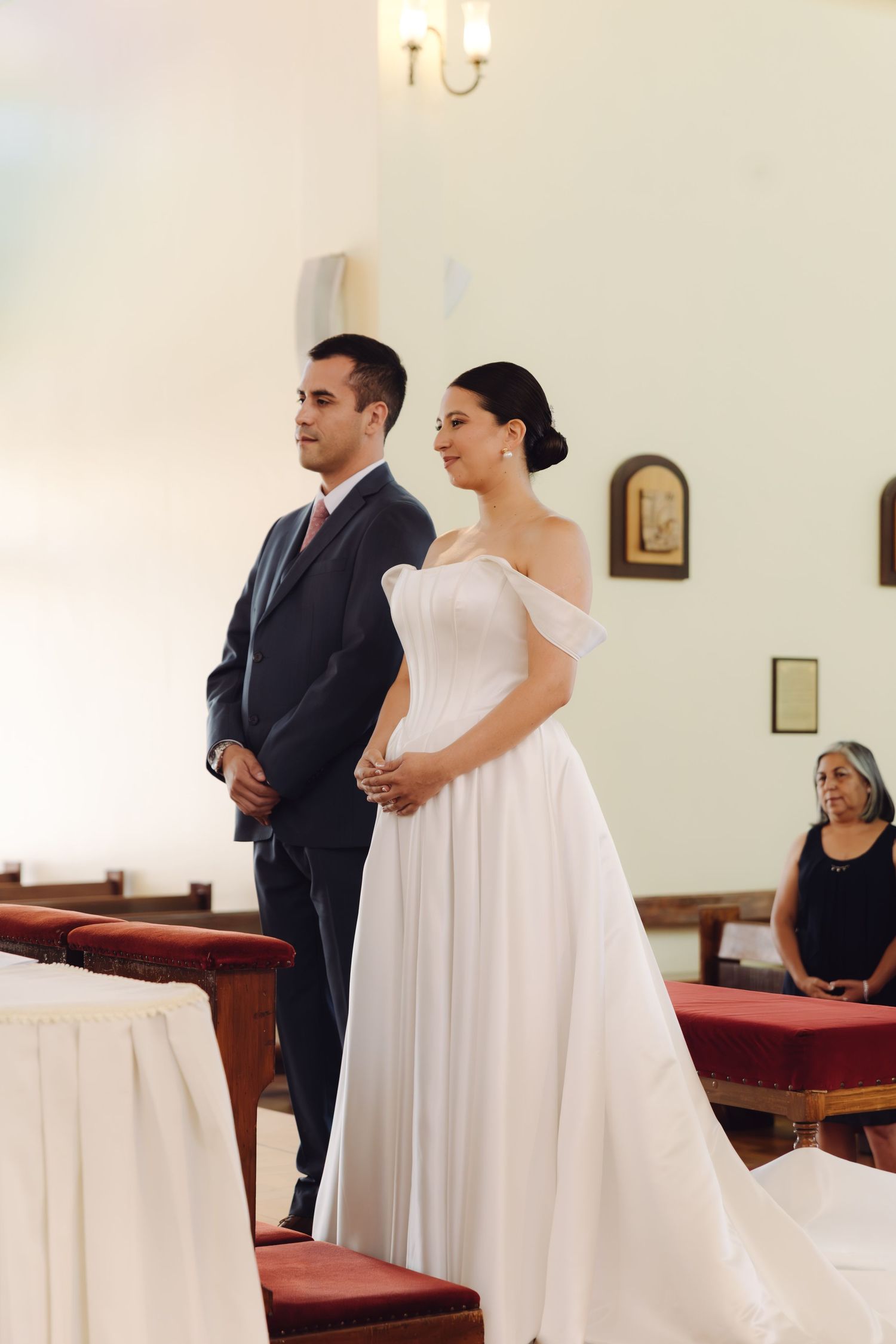 Bride in white off-shoulder gown and groom in navy suit stand together during church wedding ceremony.