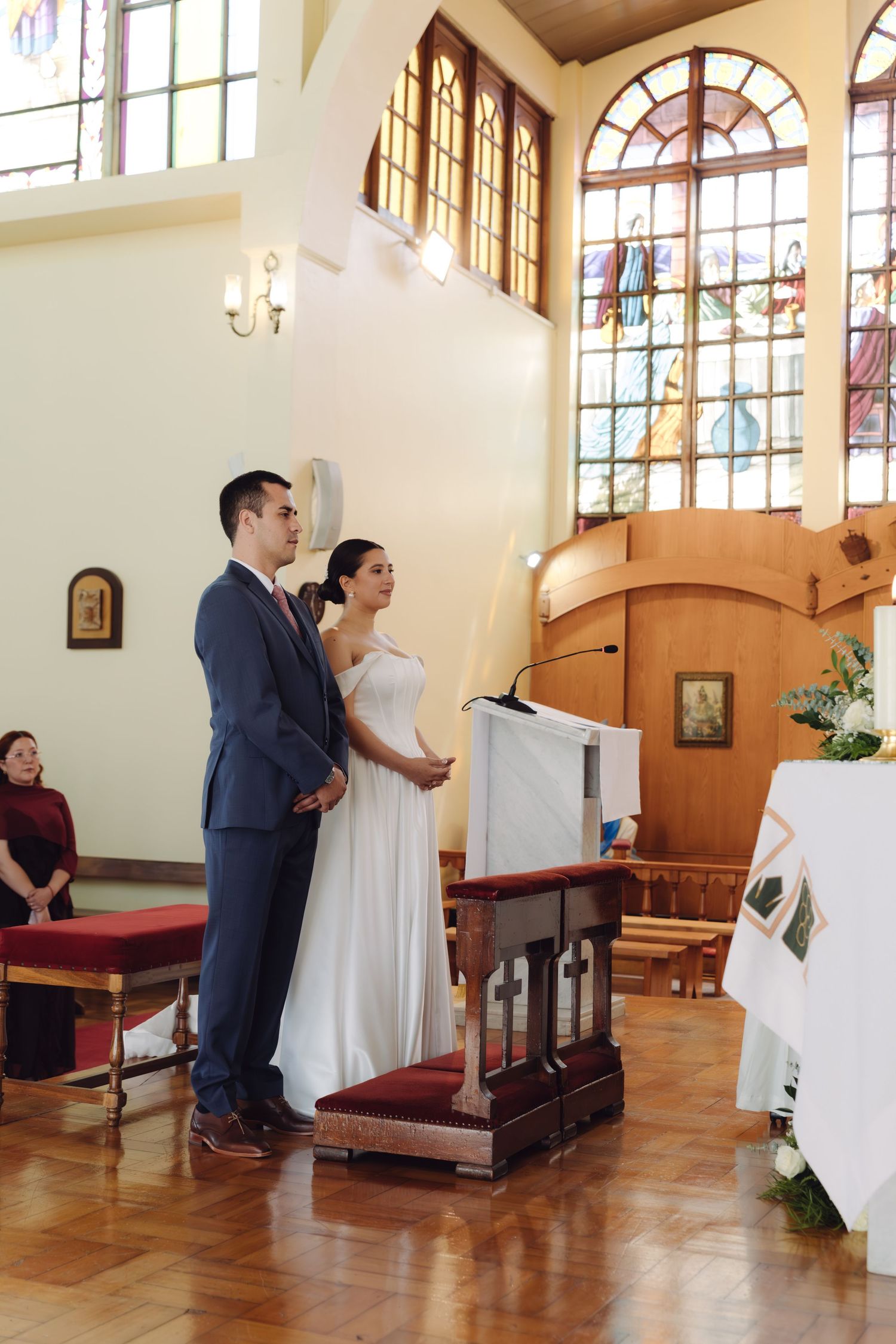 Couple stands at church altar with stained glass windows and floral arrangements during wedding ceremony.
