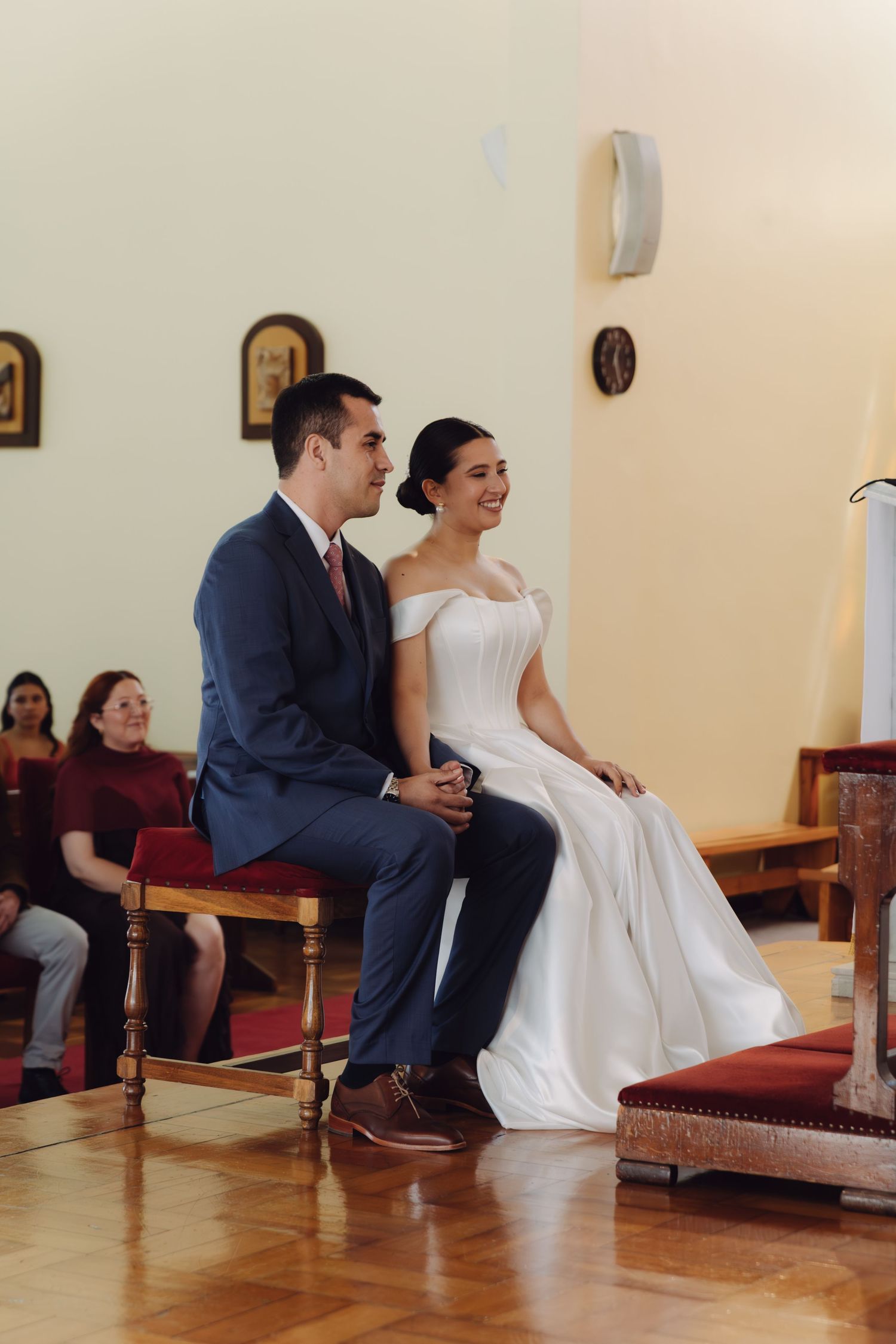 Smiling bride and groom sit together on wooden chairs during a joyful church wedding ceremony.