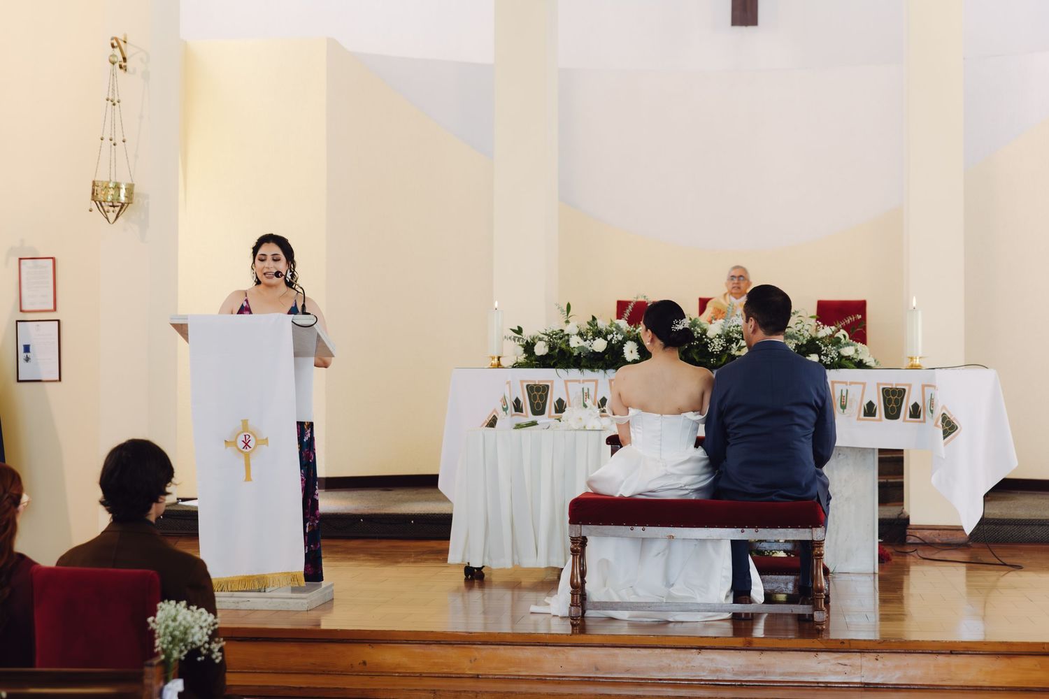 Wedding guest reads at the podium while bride and groom sit at a flower-decorated altar.
