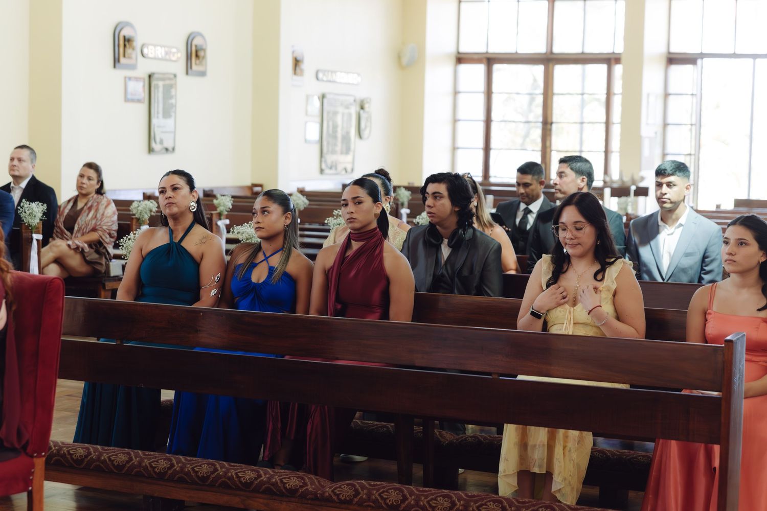 Elegantly dressed wedding guests in colorful attire sit in church pews during a wedding ceremony.
