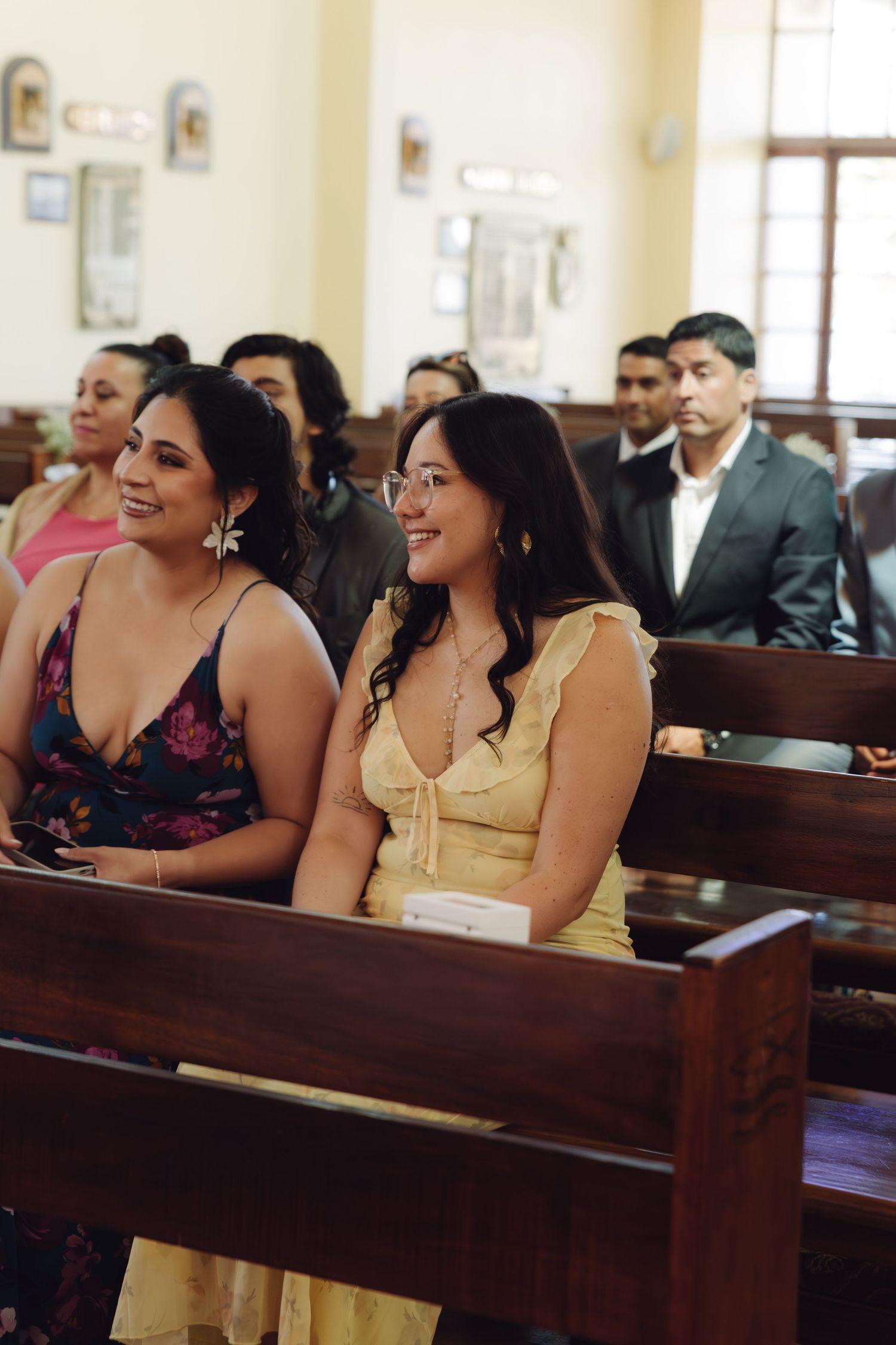 Two smiling female wedding guests in floral and yellow dresses sit in church pews during ceremony.