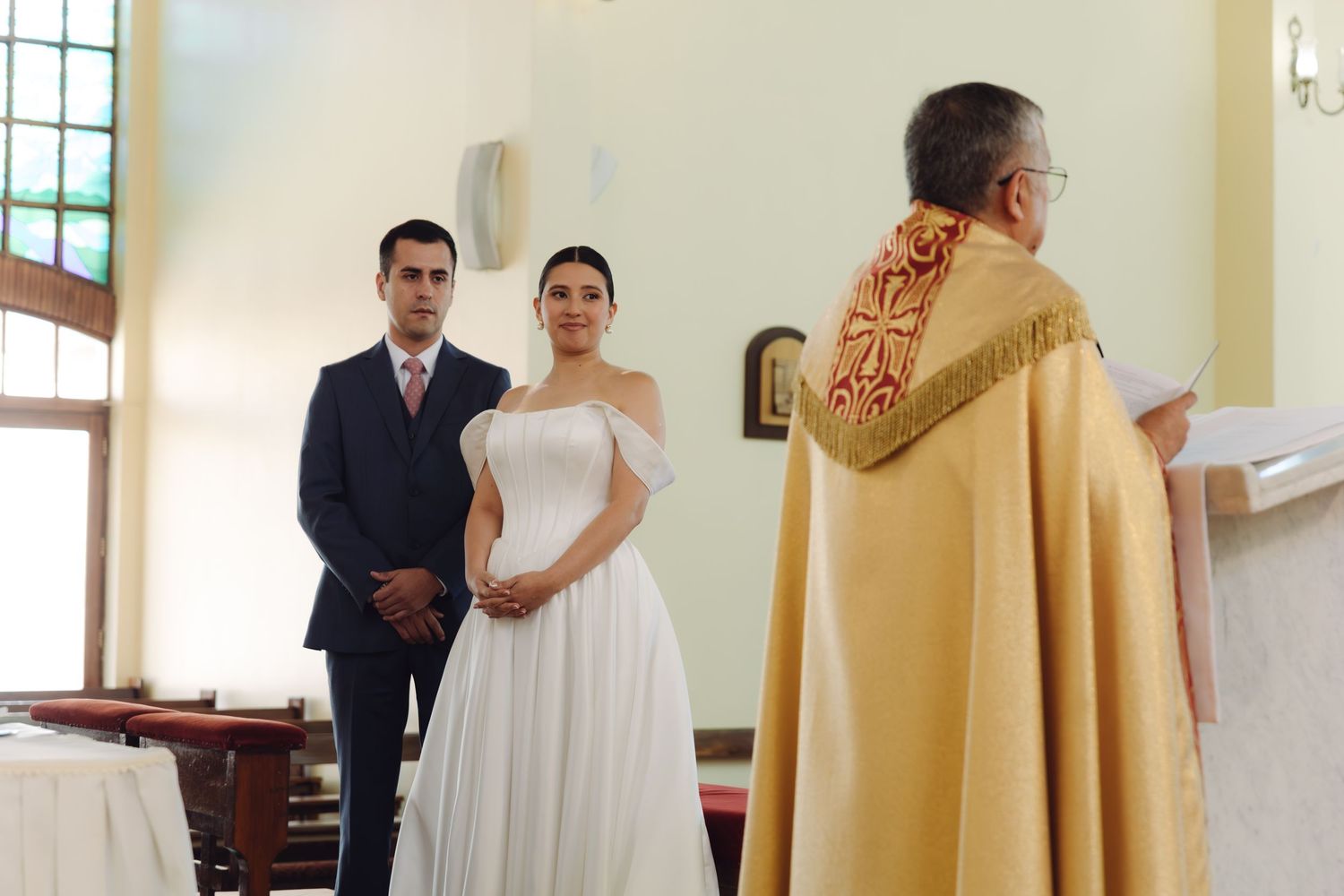 Bride and groom stand facing a priest in golden vestments during a traditional church wedding ceremony.