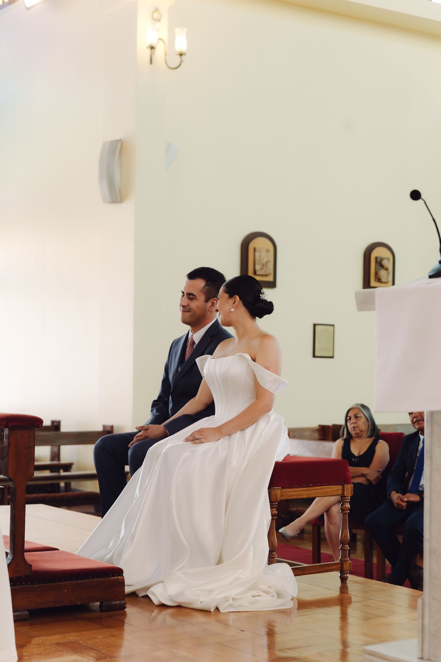 Bride in white off-shoulder gown and groom in navy suit seated together during church wedding ceremony.