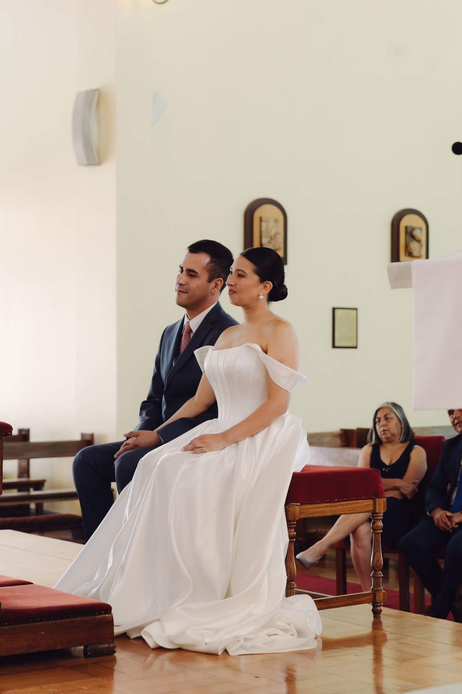 Elegant bride and groom sitting at church altar during wedding ceremony with guests in background.