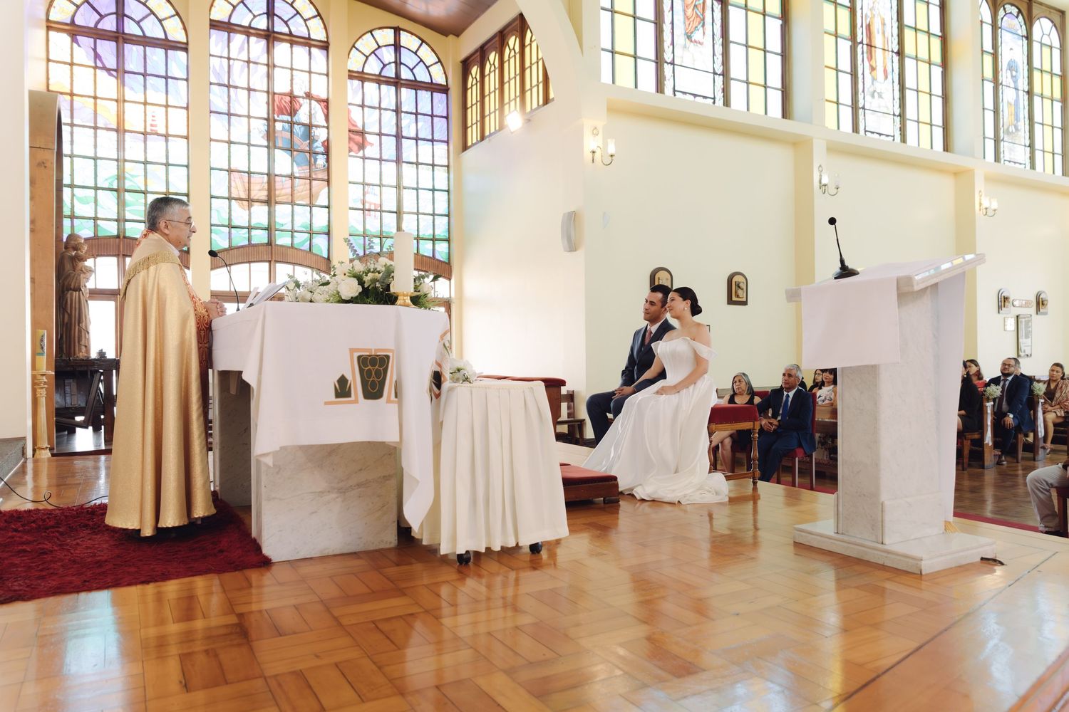 Priest officiating wedding ceremony as bride and groom sit at the altar of a church.