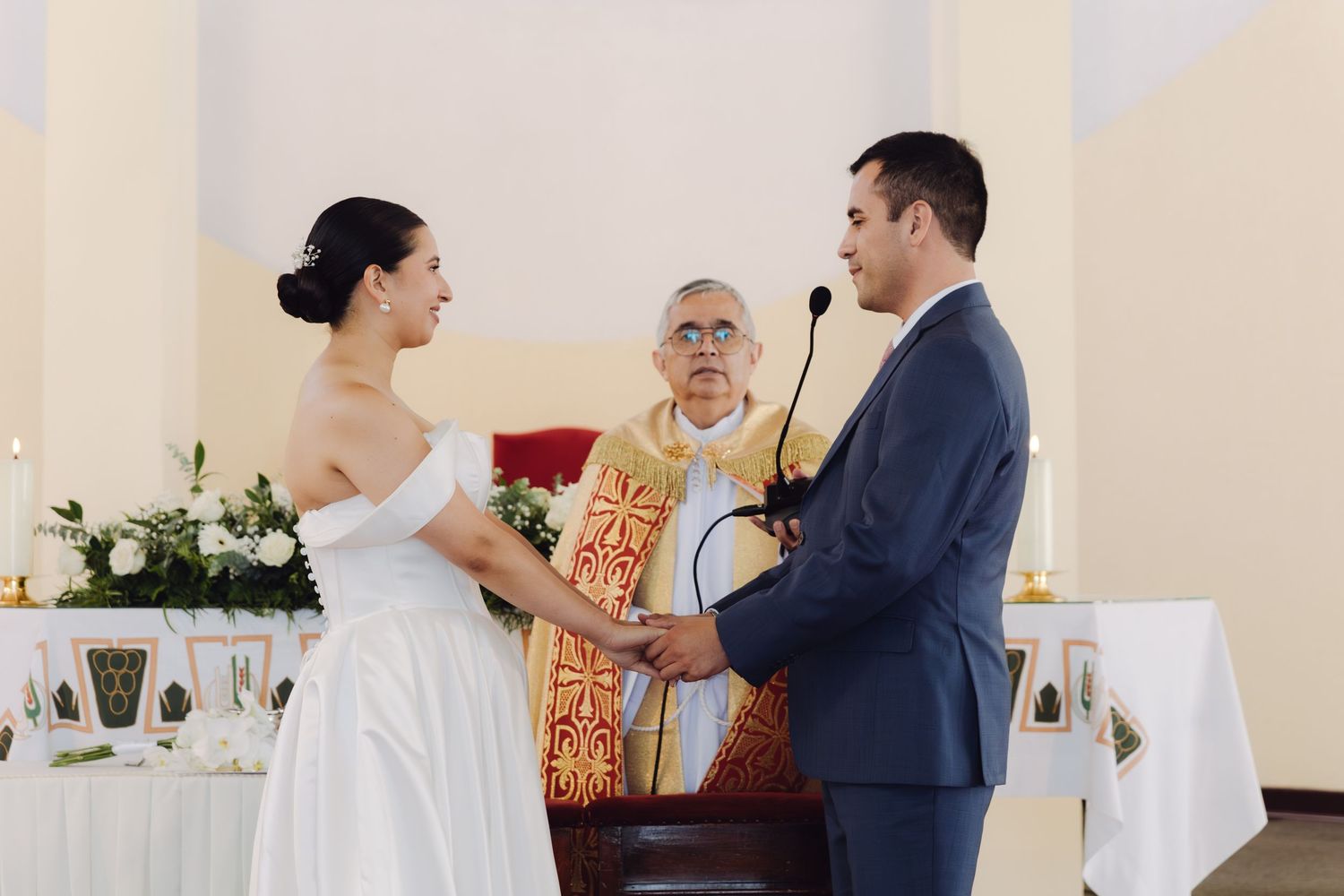 Bride and groom exchanging vows while holding hands before priest at church wedding ceremony.