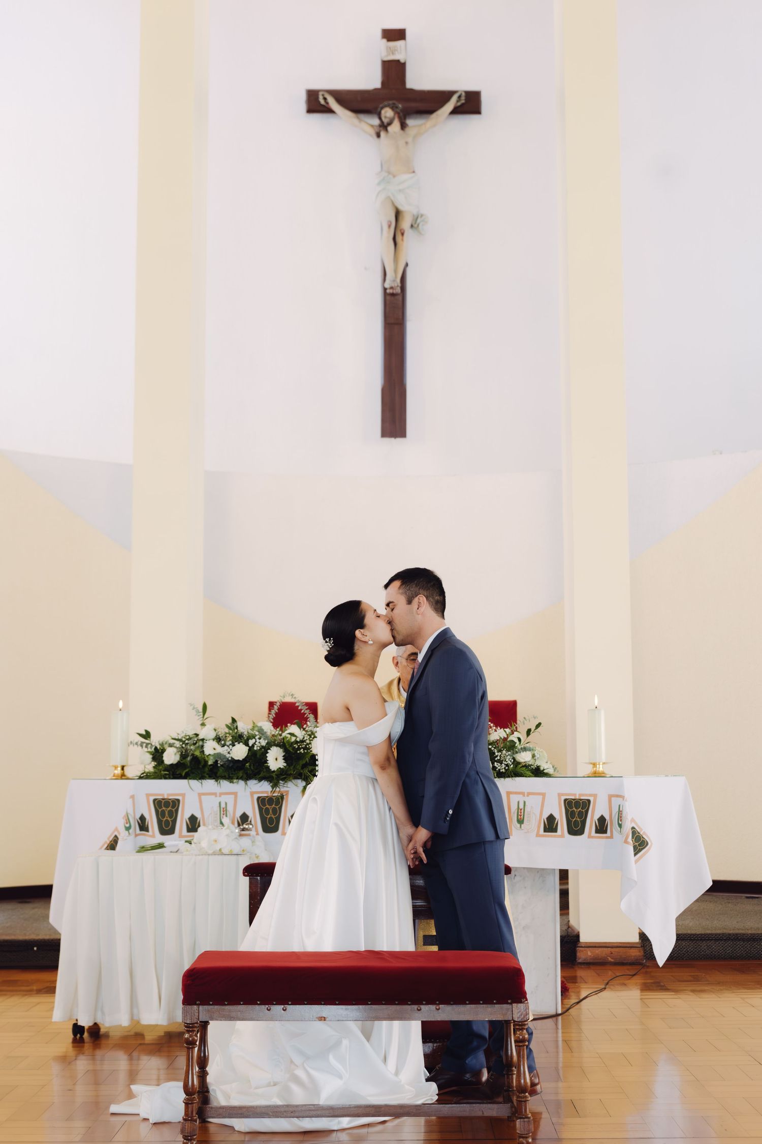 Bride and groom sharing first kiss at church altar beneath crucifix after wedding ceremony.