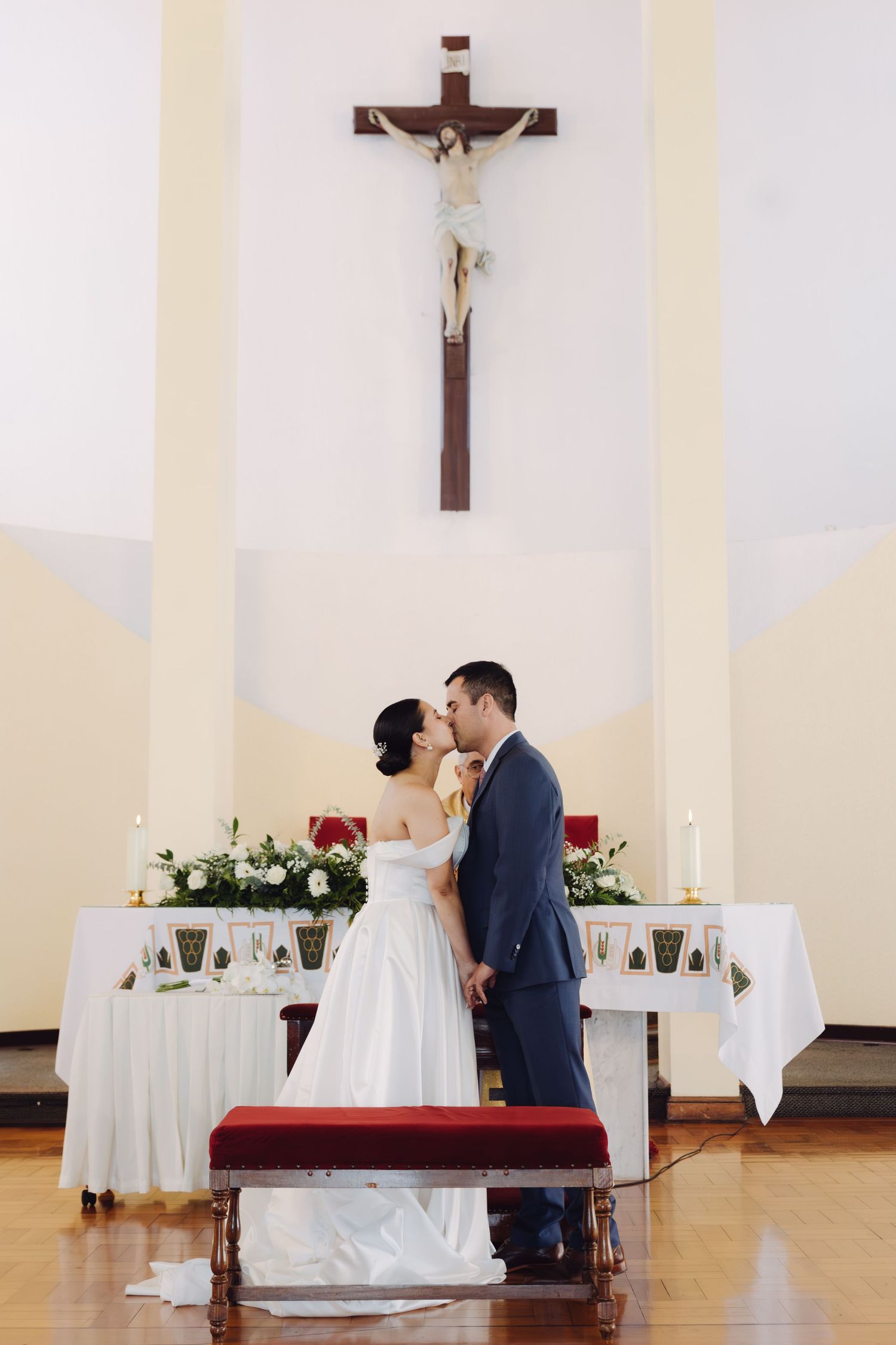 Newlyweds kissing at church altar with crucifix above and white floral arrangements on either side.