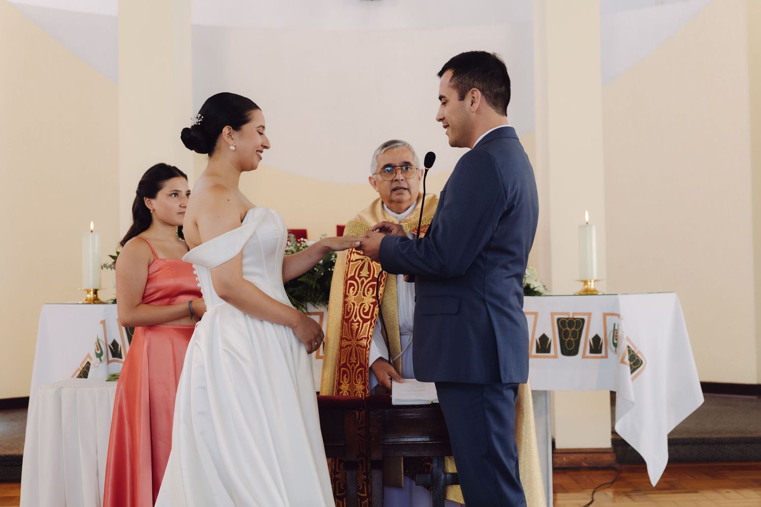Bride and groom exchanging rings during church wedding ceremony with bridesmaid and priest present.