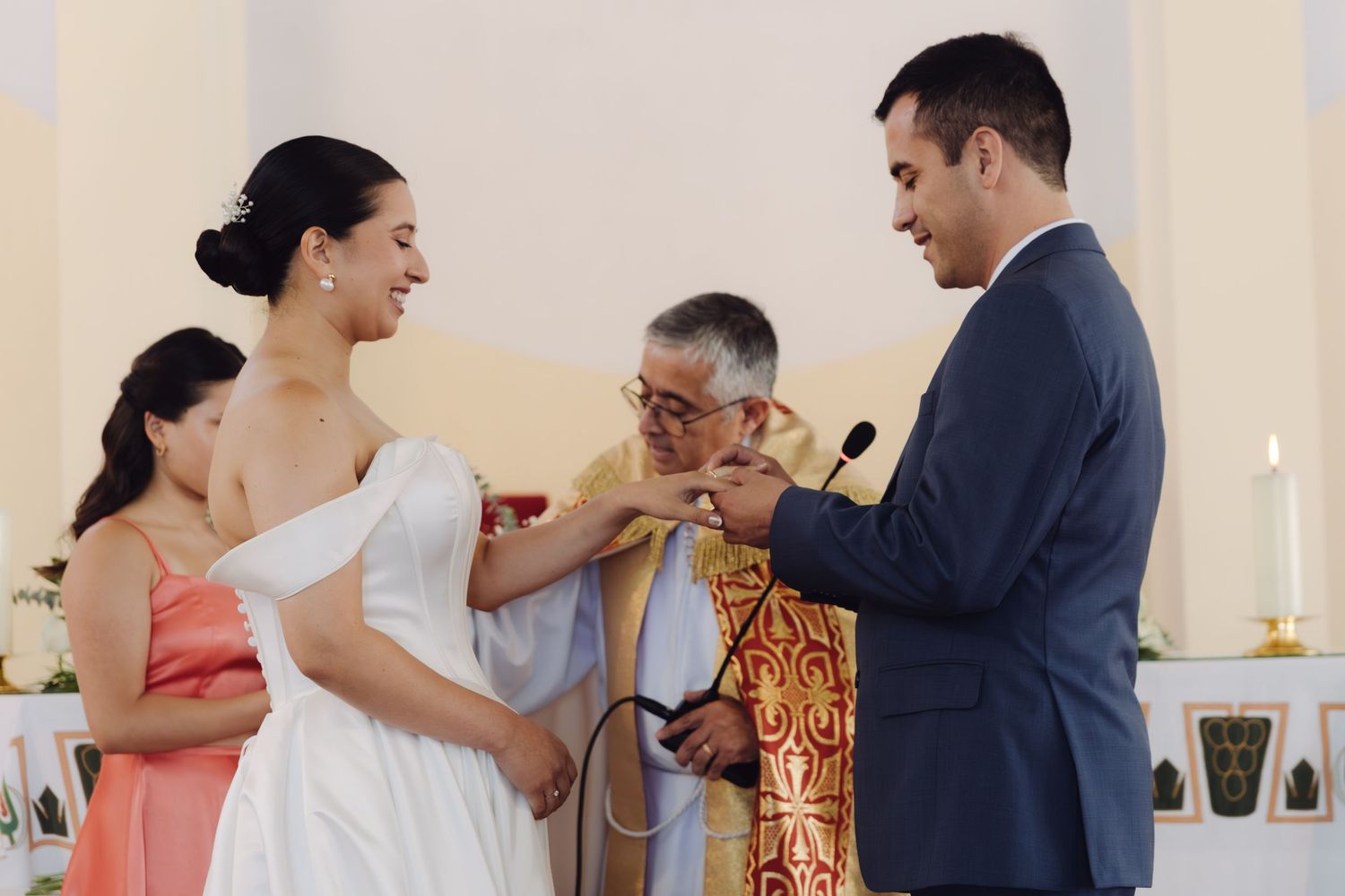 Priest officiates wedding ring exchange ceremony with bride in white off-shoulder gown and groom in navy suit.