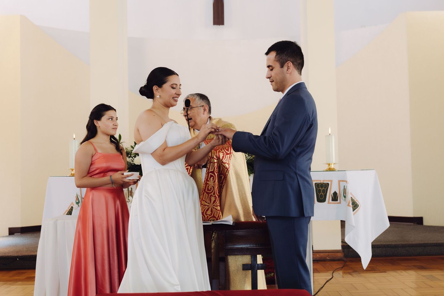 Bride and groom exchange wedding rings at church altar with bridesmaid and officiant present.