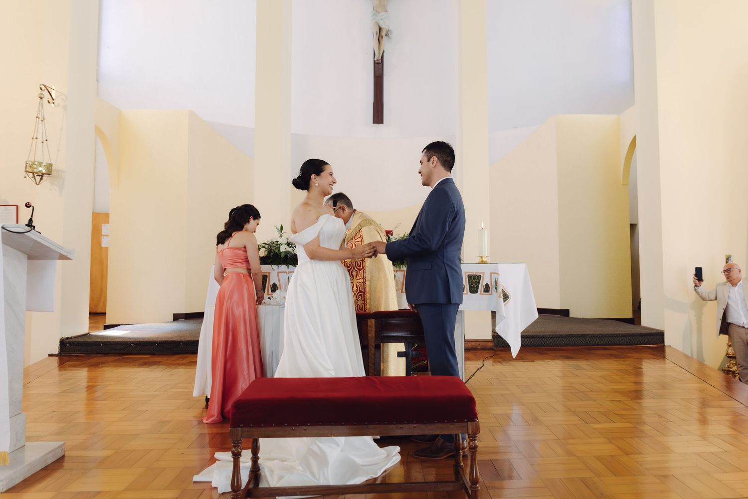 Couple stands at church altar during wedding ceremony with bridesmaid in coral dress and wooden crucifix.