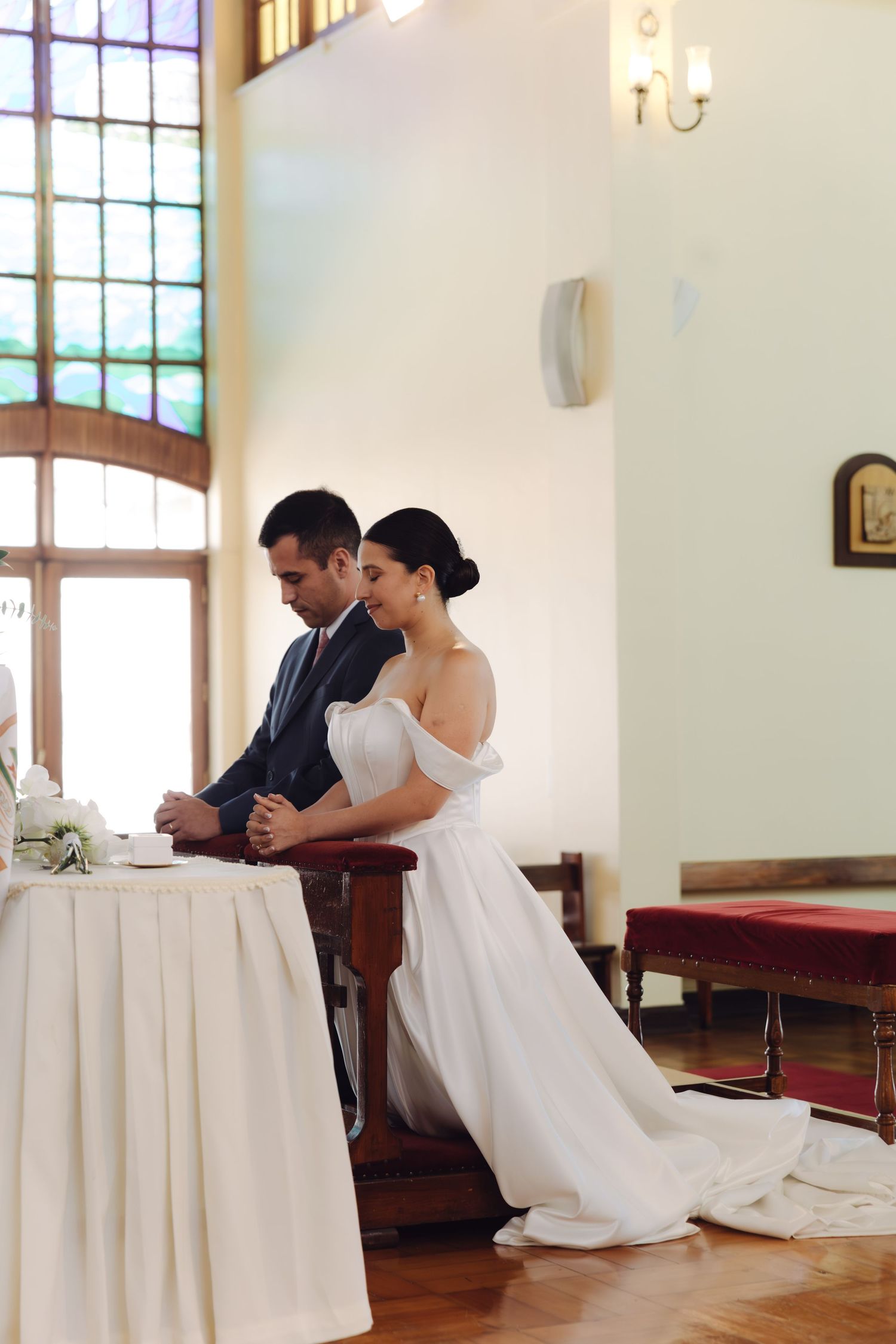 Bride and groom kneel in prayer at church pew during wedding ceremony beside stained glass window.