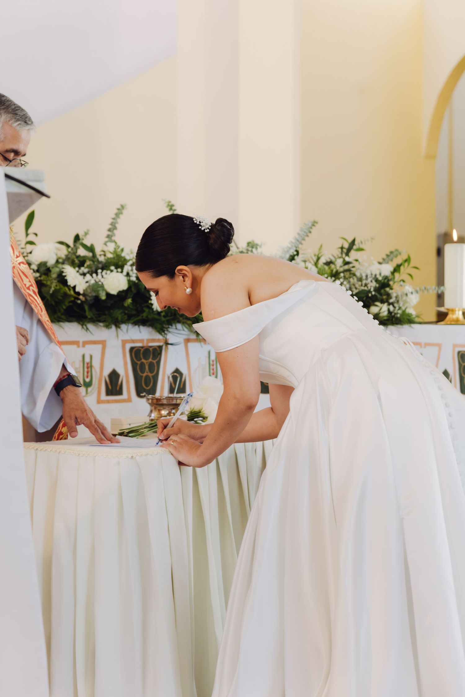 Bride in off-shoulder white gown signs wedding register at flower-adorned altar during church ceremony.