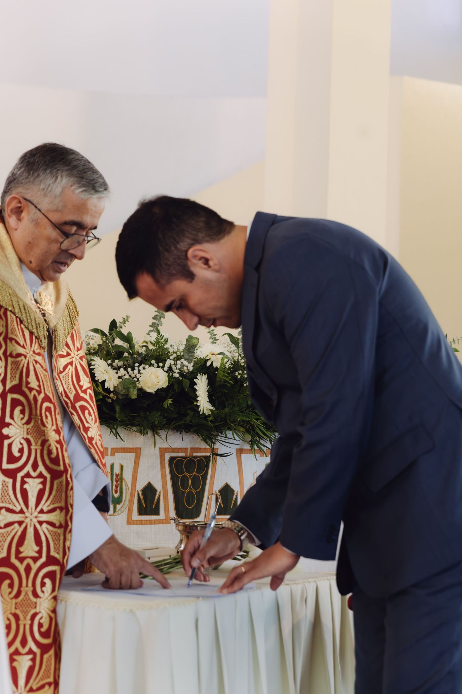Groom in navy suit signs official wedding register document beside priest in gold ceremonial vestments.
