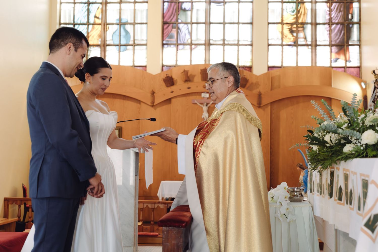 Priest in gold vestments presents wedding certificate to couple standing before stained glass church windows.