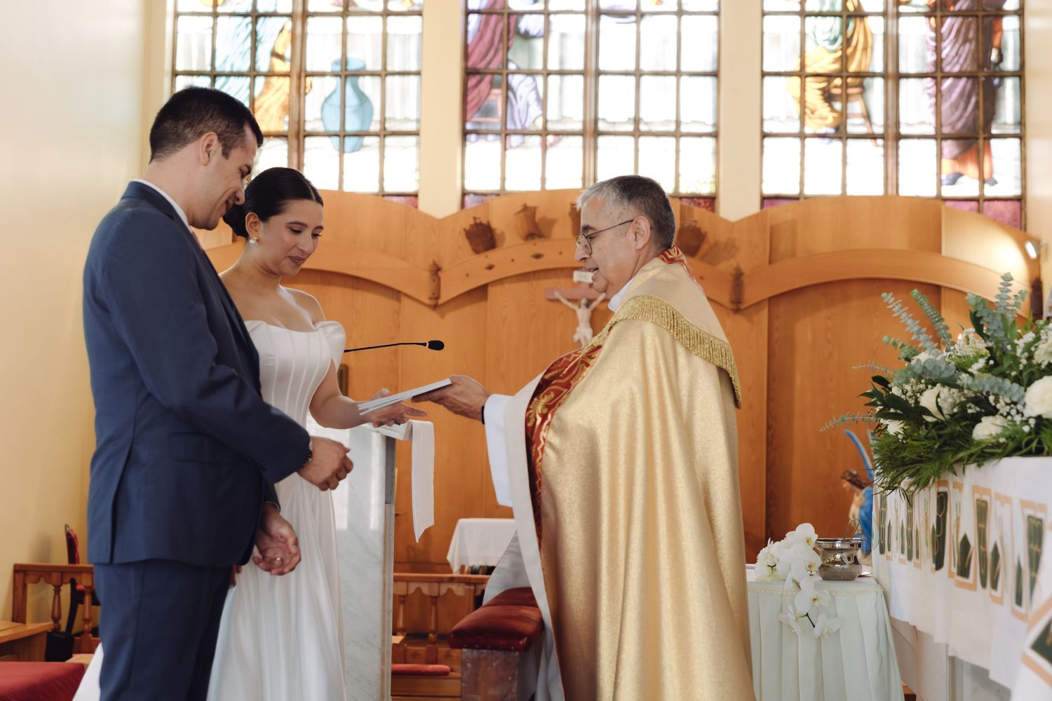 Officiant in gold robes reads from document to bride and groom during church wedding ceremony.