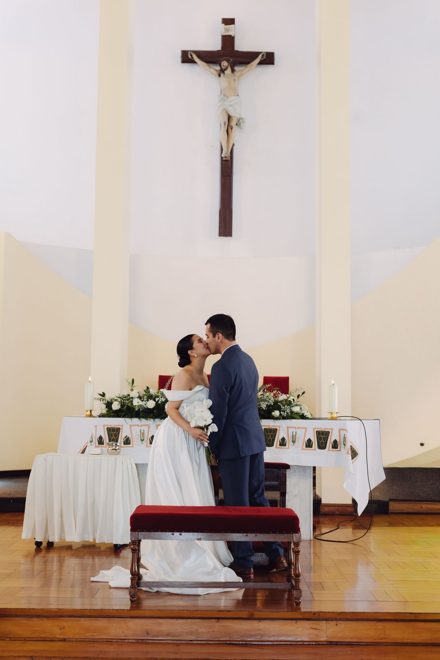 Newlyweds share first kiss at church altar beneath crucifix after Catholic wedding ceremony.