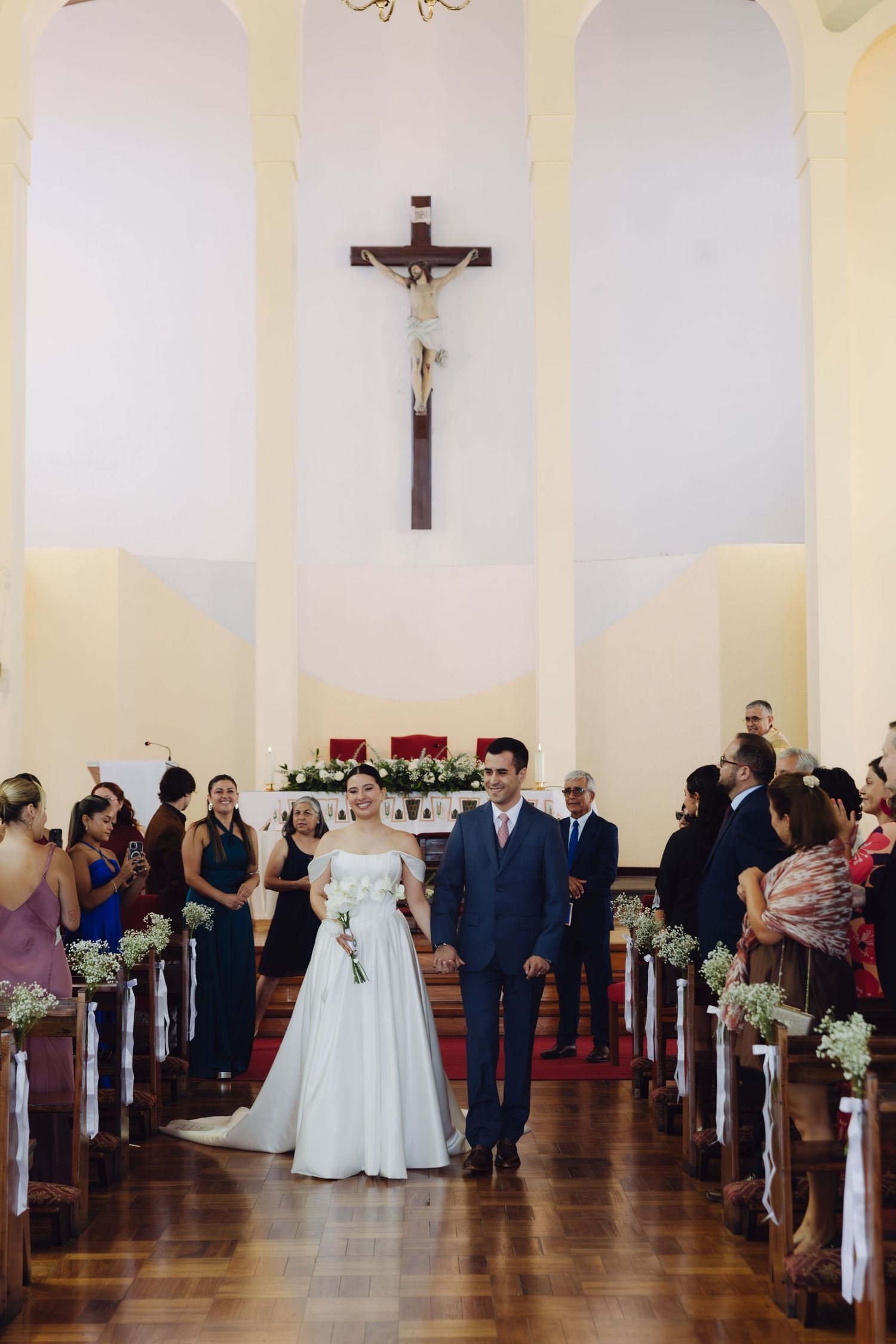 Bride and groom hold hands walking down the aisle of a Catholic church with crucifix.