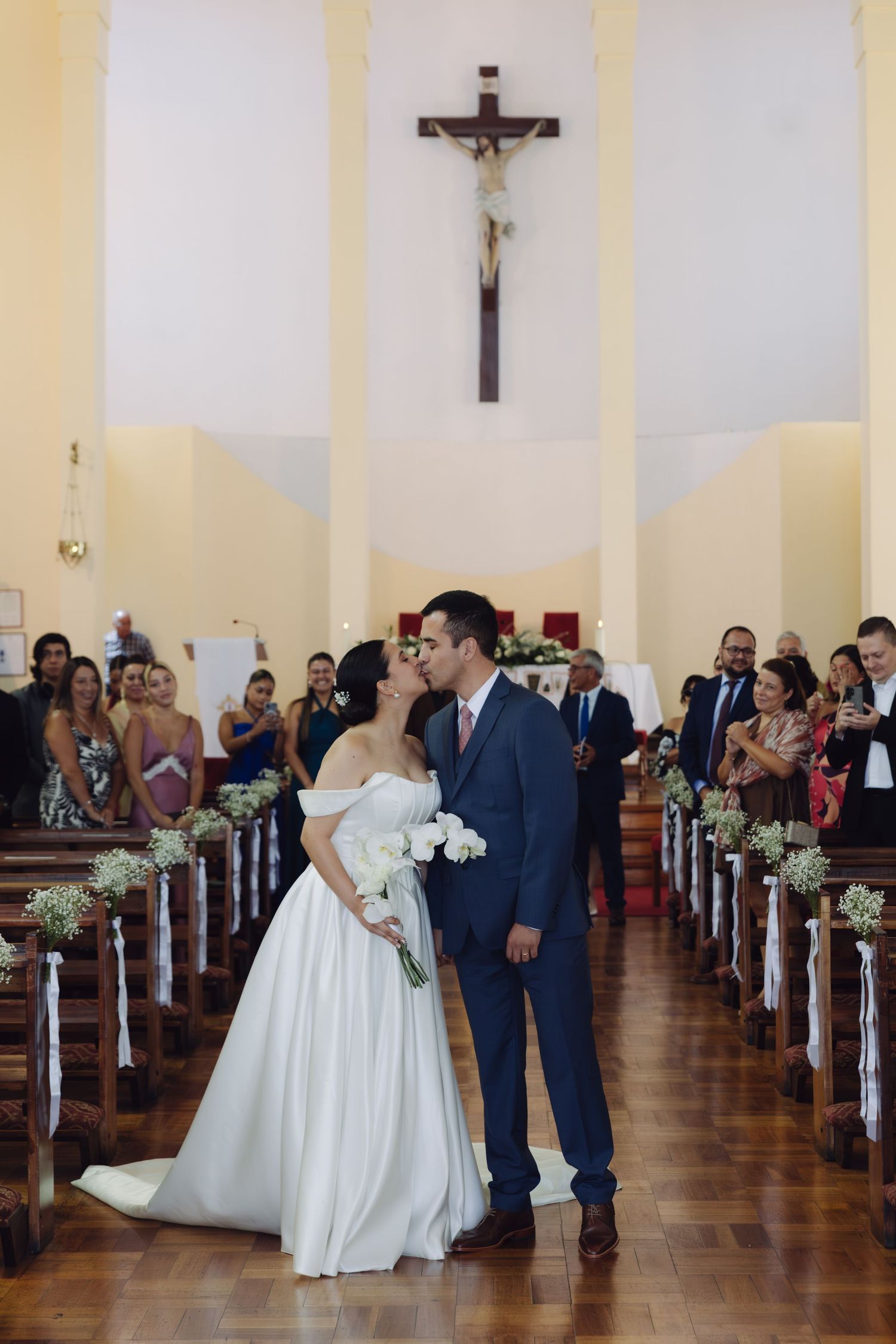 Newlyweds kiss in the center aisle of a Catholic church with wooden pews.