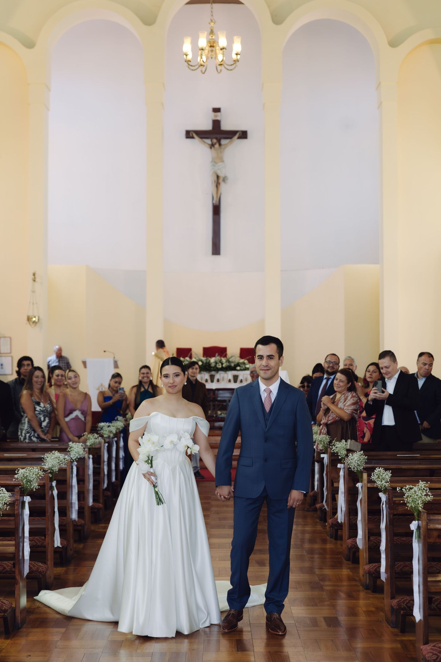 Bride holding white bouquet and groom walk hand in hand down the church aisle.
