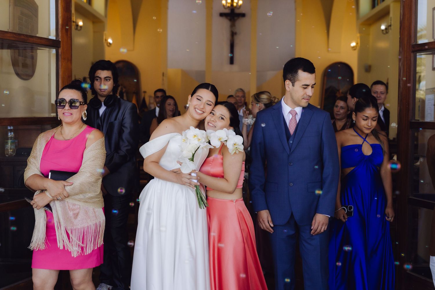 Bride in white gown stands with wedding party guests at church entrance.