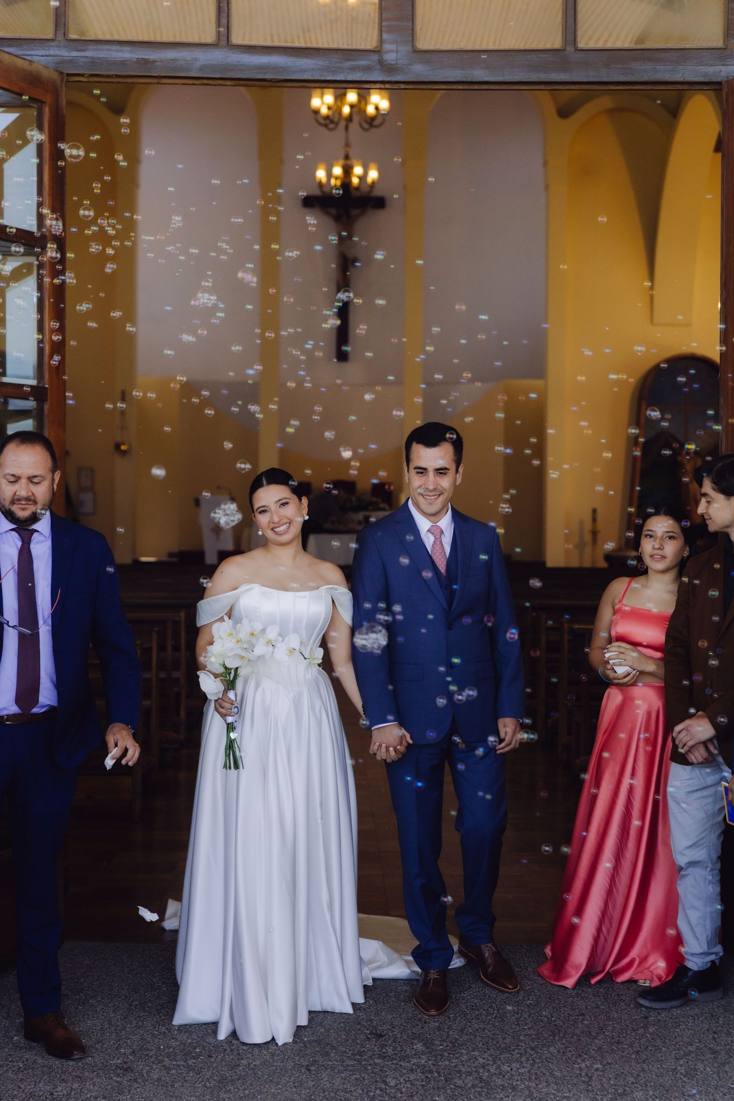 Bride and groom exit church surrounded by bubbles, holding hands with guests nearby.