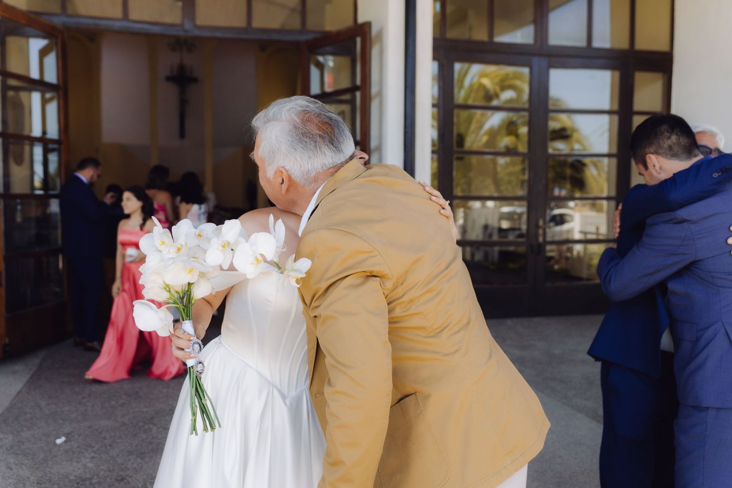 Older gentleman lovingly kisses bride holding white orchid bouquet outside the church.