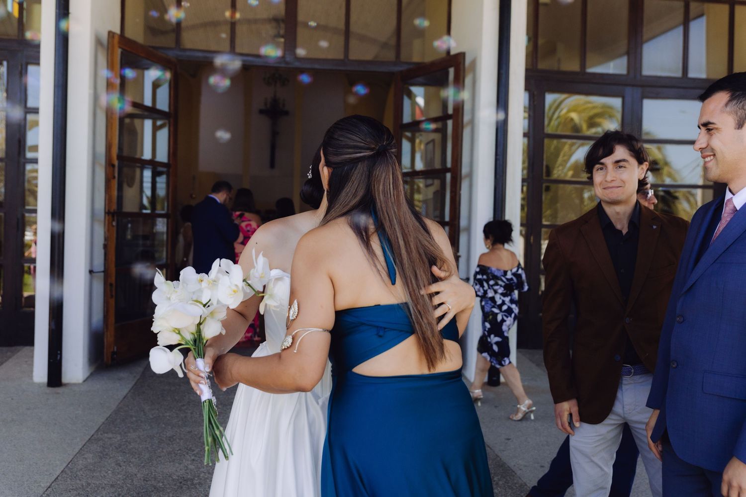 Bride holding white orchid bouquet receives warm embrace from guest outside church doors.