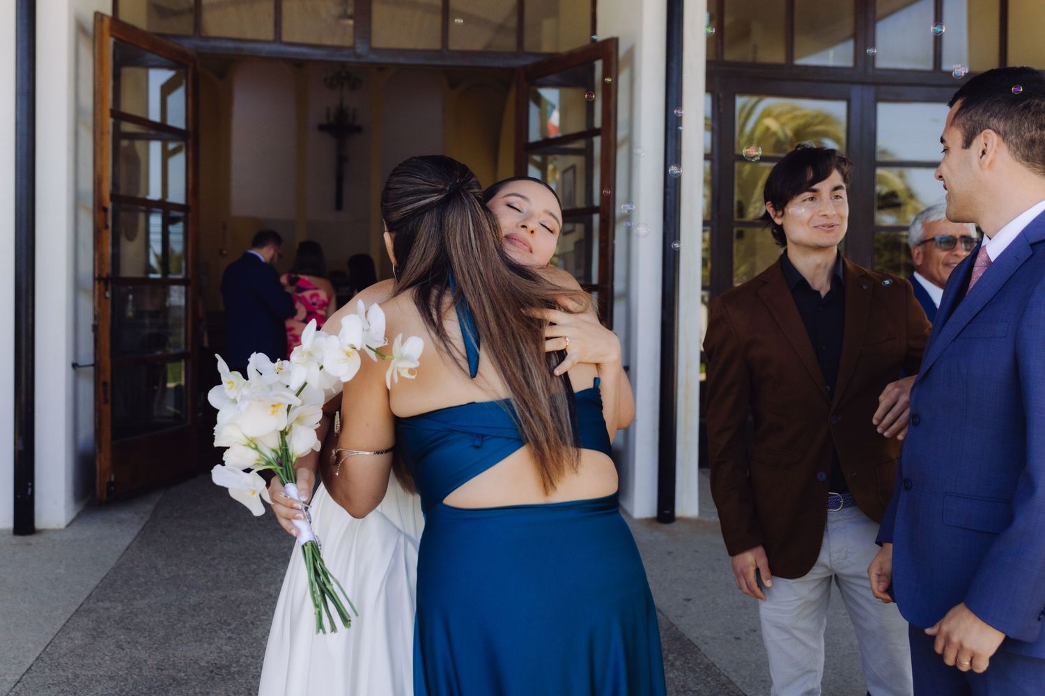 Bride in white gown hugs a guest in blue dress while holding white orchid bouquet outside.