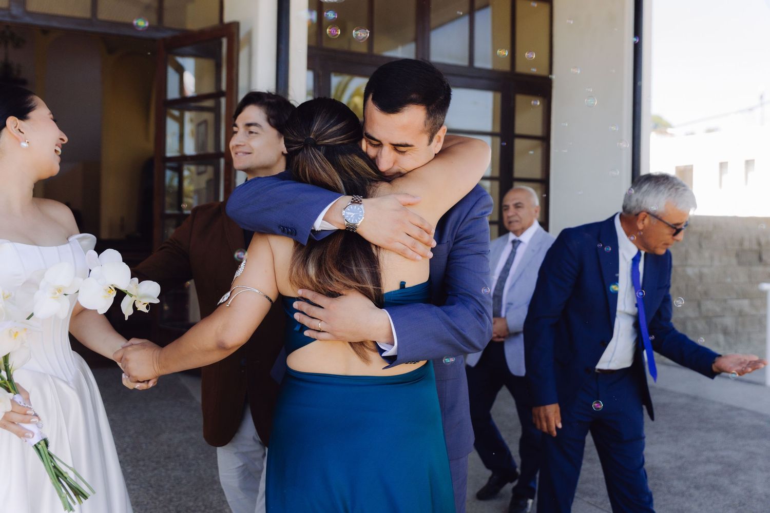 Wedding guests embracing with warm hugs outside a venue, bride holding white orchids nearby.