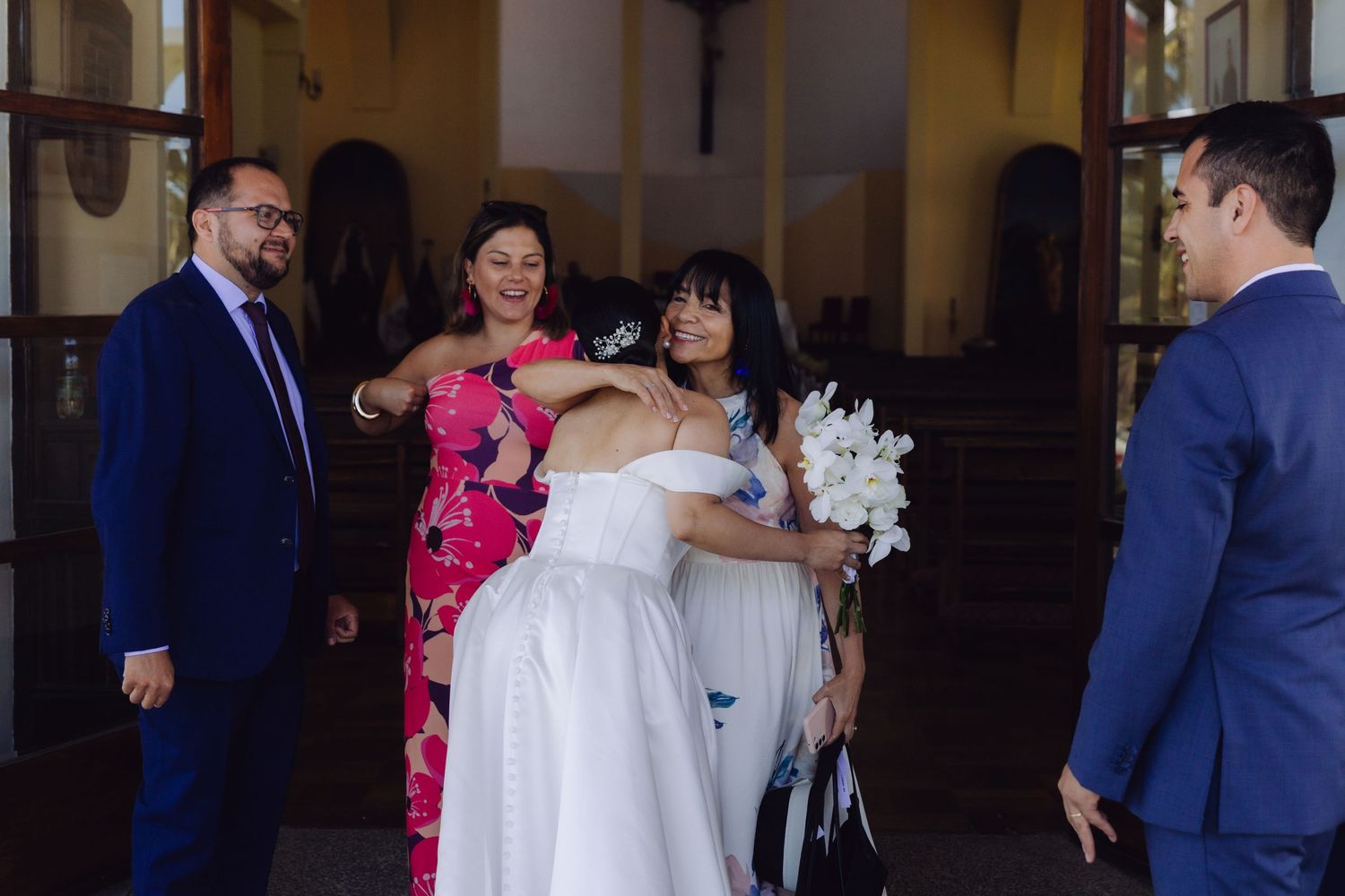 Bride in white gown holding white lilies greeted by guests outside a church entrance.