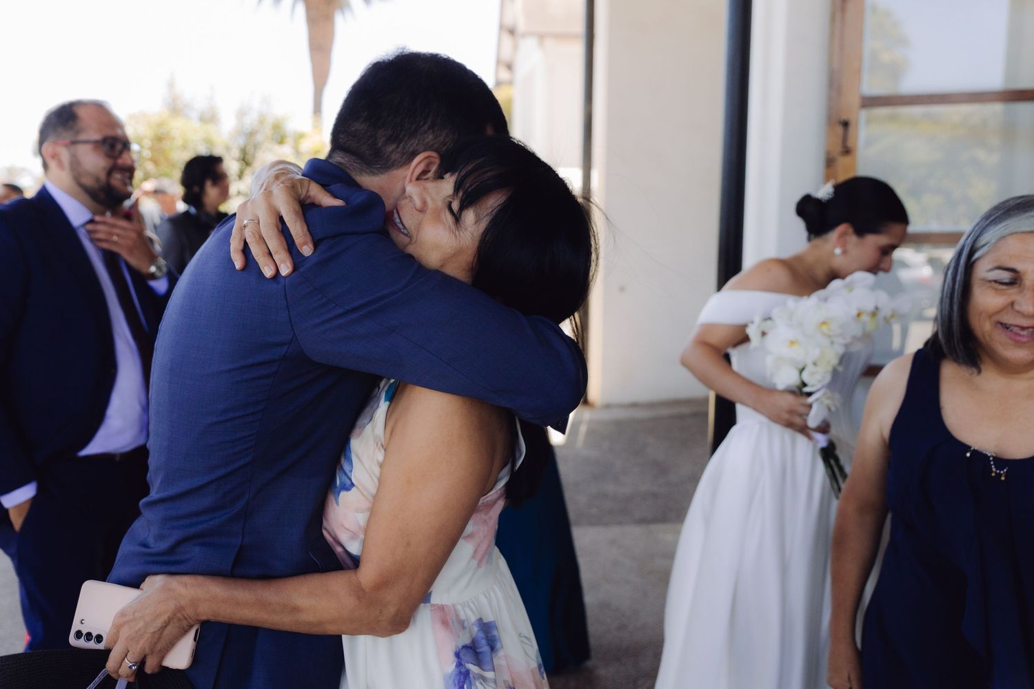 Emotional wedding guest hugging someone in a blue suit outside a ceremony venue.