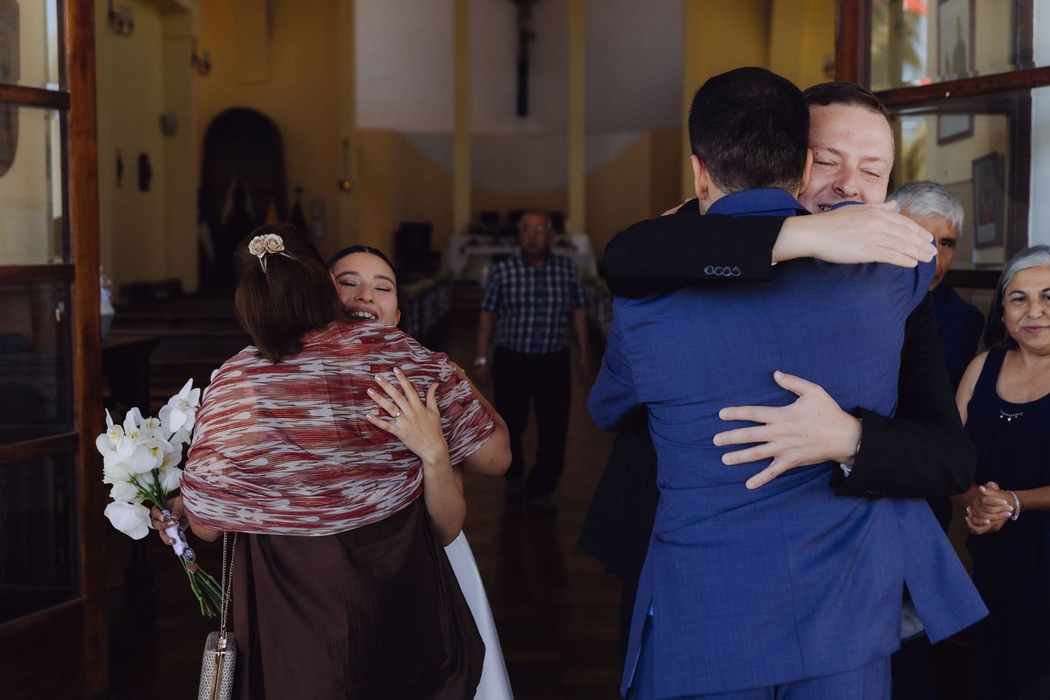 Wedding guests sharing warm embraces outside a venue, bride visible in background.